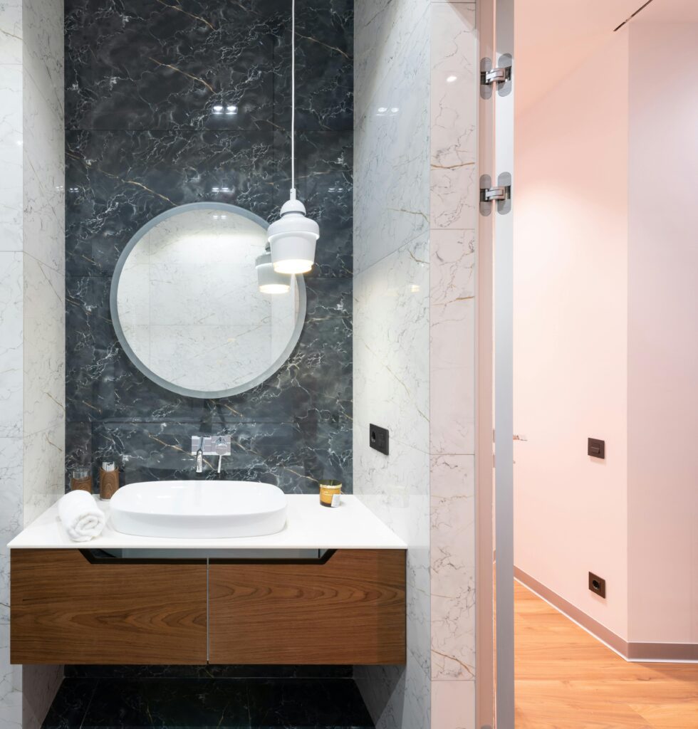 Modern bright bathroom interior with round mirror near lamp hanging from ceiling and white sink on cabinet