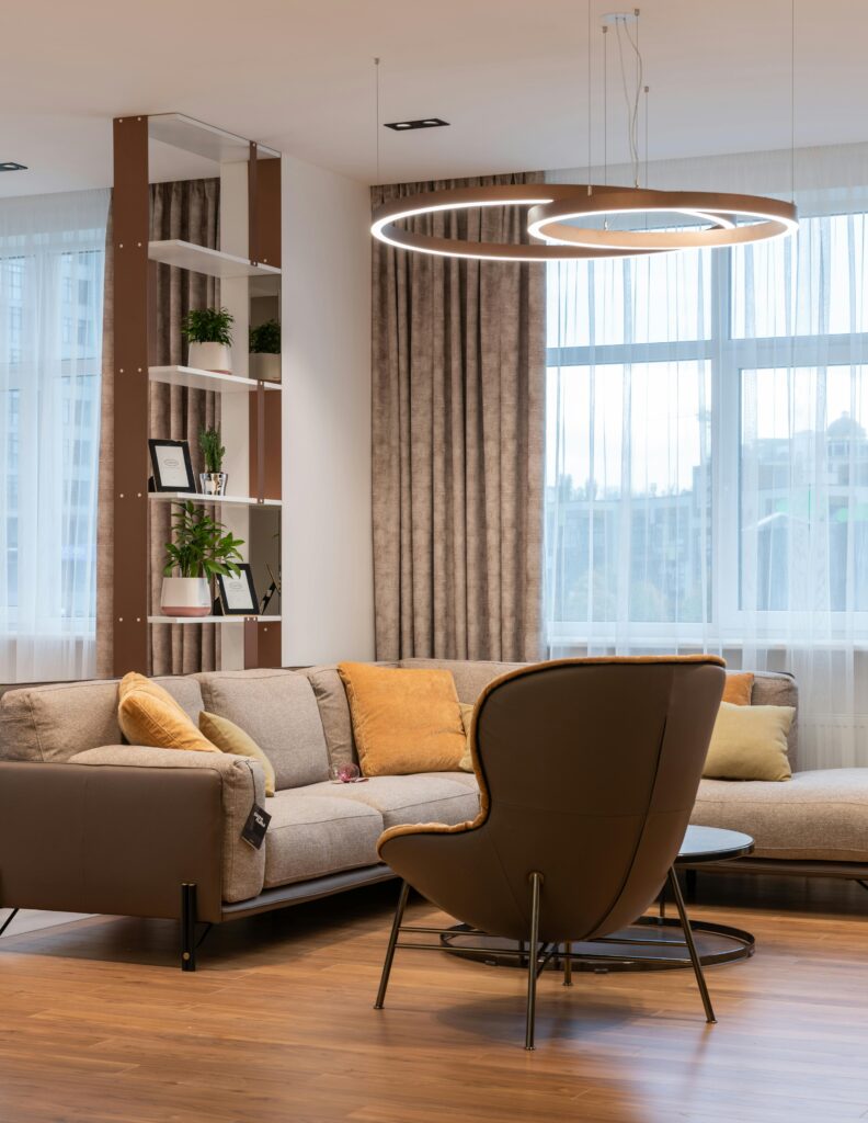 Modern living room interior with soft couch and pillows near armchair and round table near window with curtains and shelves with plants near lamp