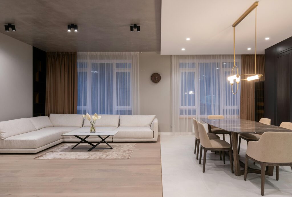 Interior of modern light flat with sofa next to dining table with chairs under lamps hanging on ceiling near windows with curtains