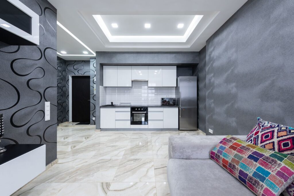 Kitchen area with white furniture placed near living room with comfortable sofa and TV in contemporary apartment with gray walls