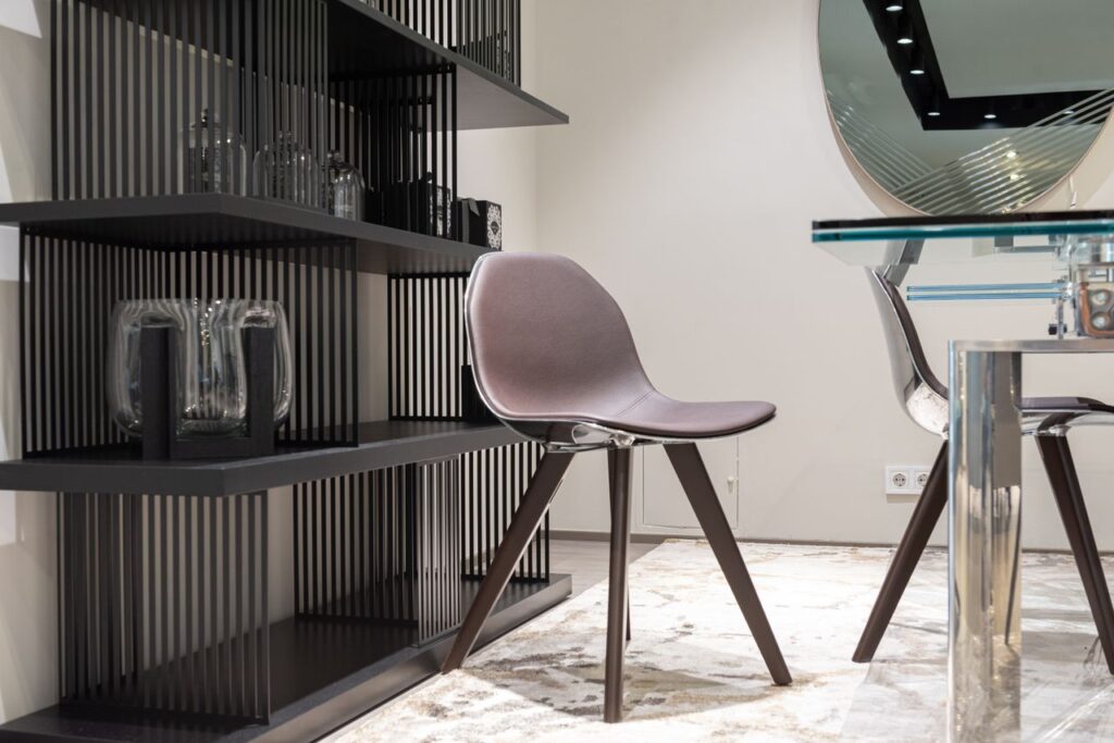 Stylish chair and table placed near shelves with glass objects and mirror hanging on wall