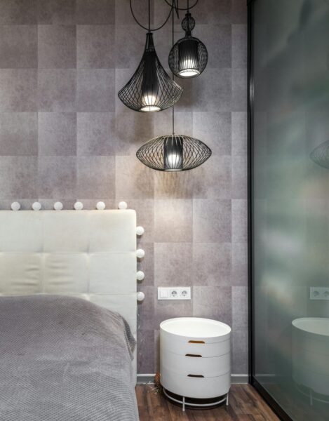 Comfortable bed with soft headboard placed at wall near white bedside table in stylish bathroom with hanging lamps in apartment
