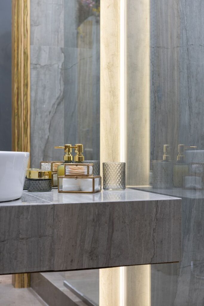 Stylish washroom with gray tiled walls and different perfume placed on marble counter near sink with reflective mirror at home