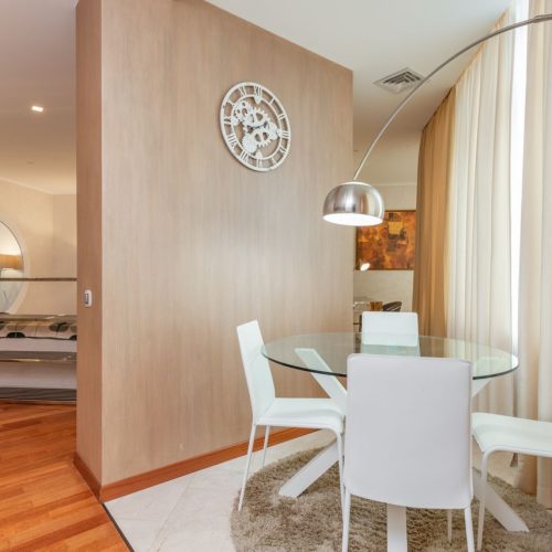 Dining zone with glass round table and white chairs separated from living room with wooden wall decorated with creative clock in daylight, distribuzione spazi interni casa